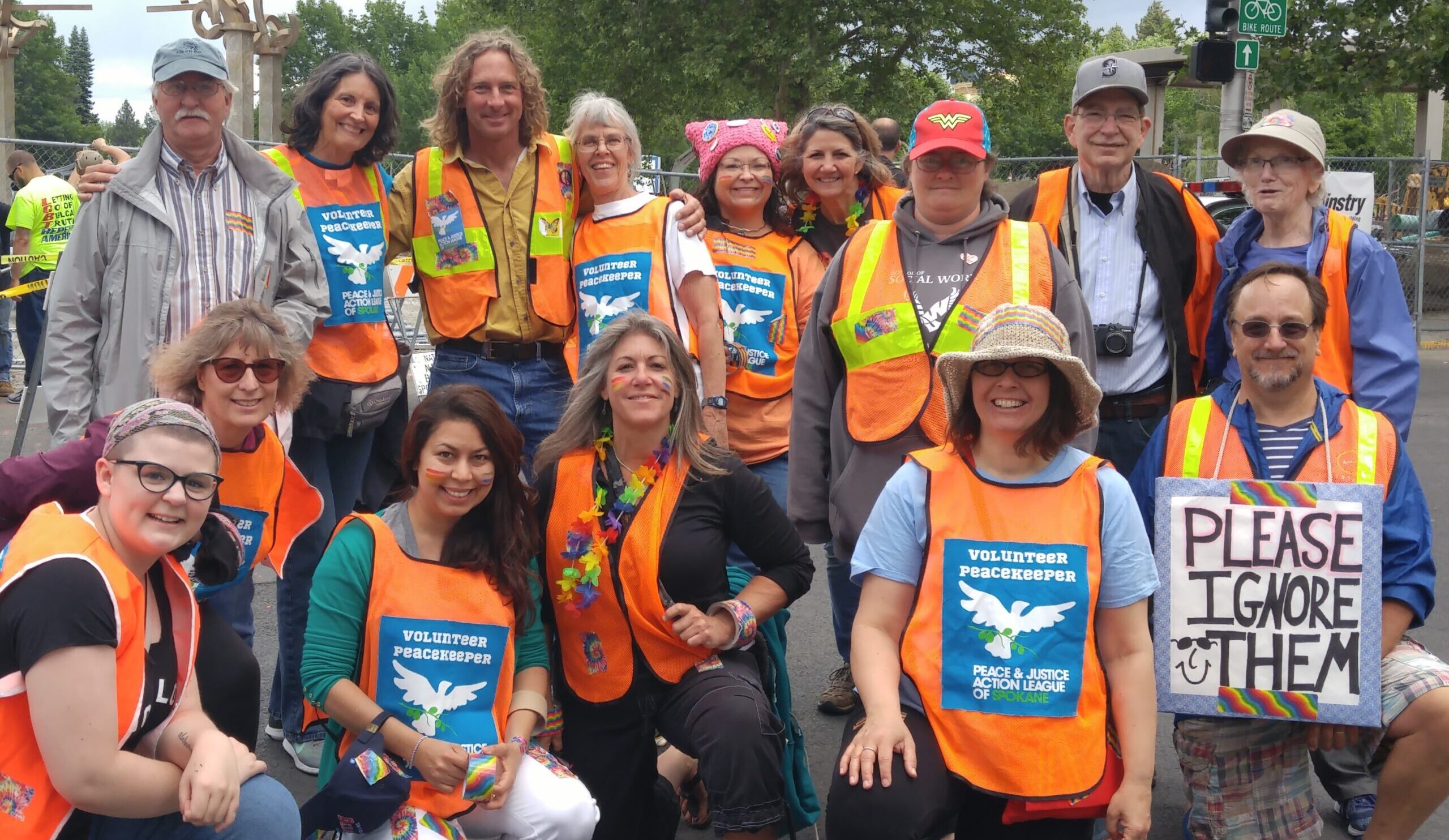 Photo of a group of 15 people wearing bright orange vests with a blue logo that reads "Peace and Justice Action League Peacekeepers"