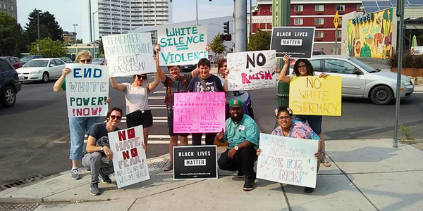 image of 9 people standing in front of a crosswalk and holding signs up to the camera. Signs read messages like, "Black Lives Matter", "End White Power Now", "No More White Supremacy", and others.