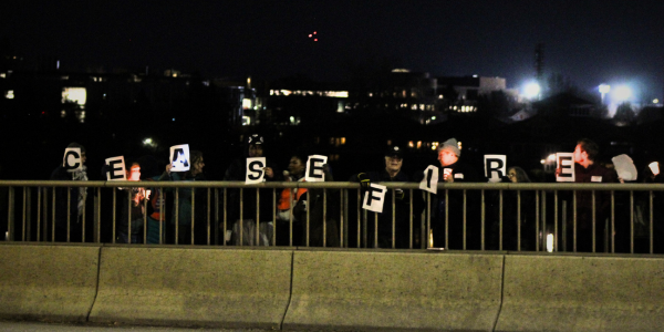 photo of 9 people standing on a bridge at nightfall. each of them are holding a sign with one letter on it. They are all standing in line to spell out "ceasefire"