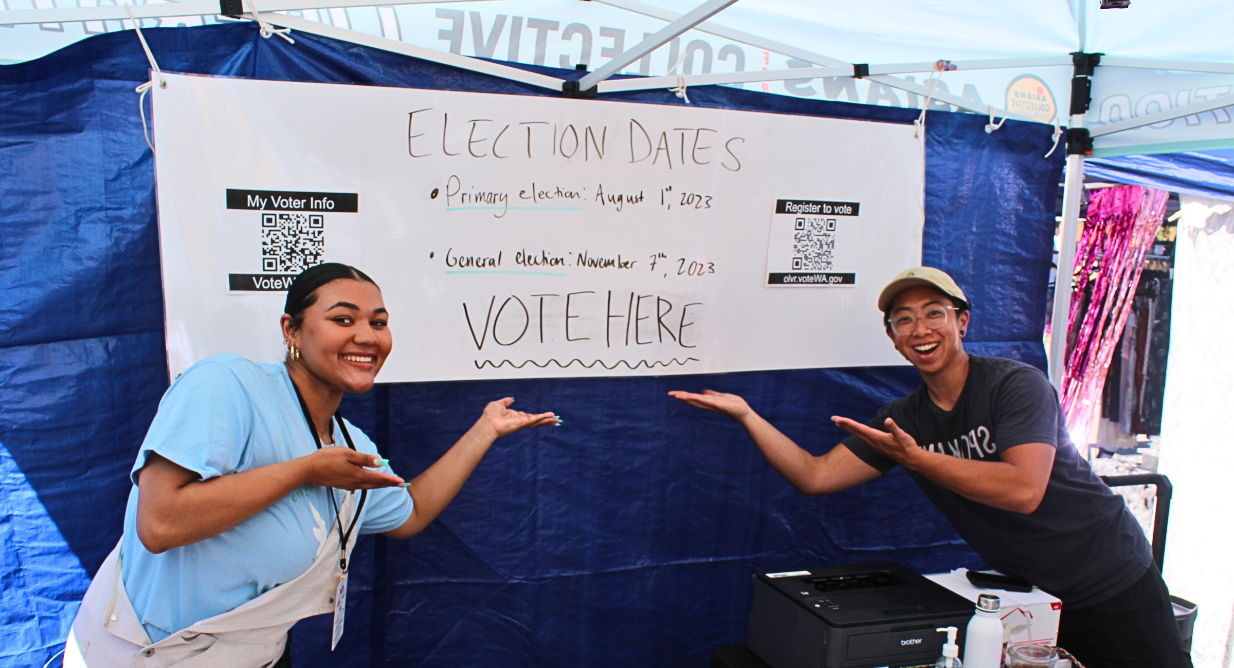 photograph of 2 people standing in front of a "get out the vote" poster and pointing to the "vote here" sign